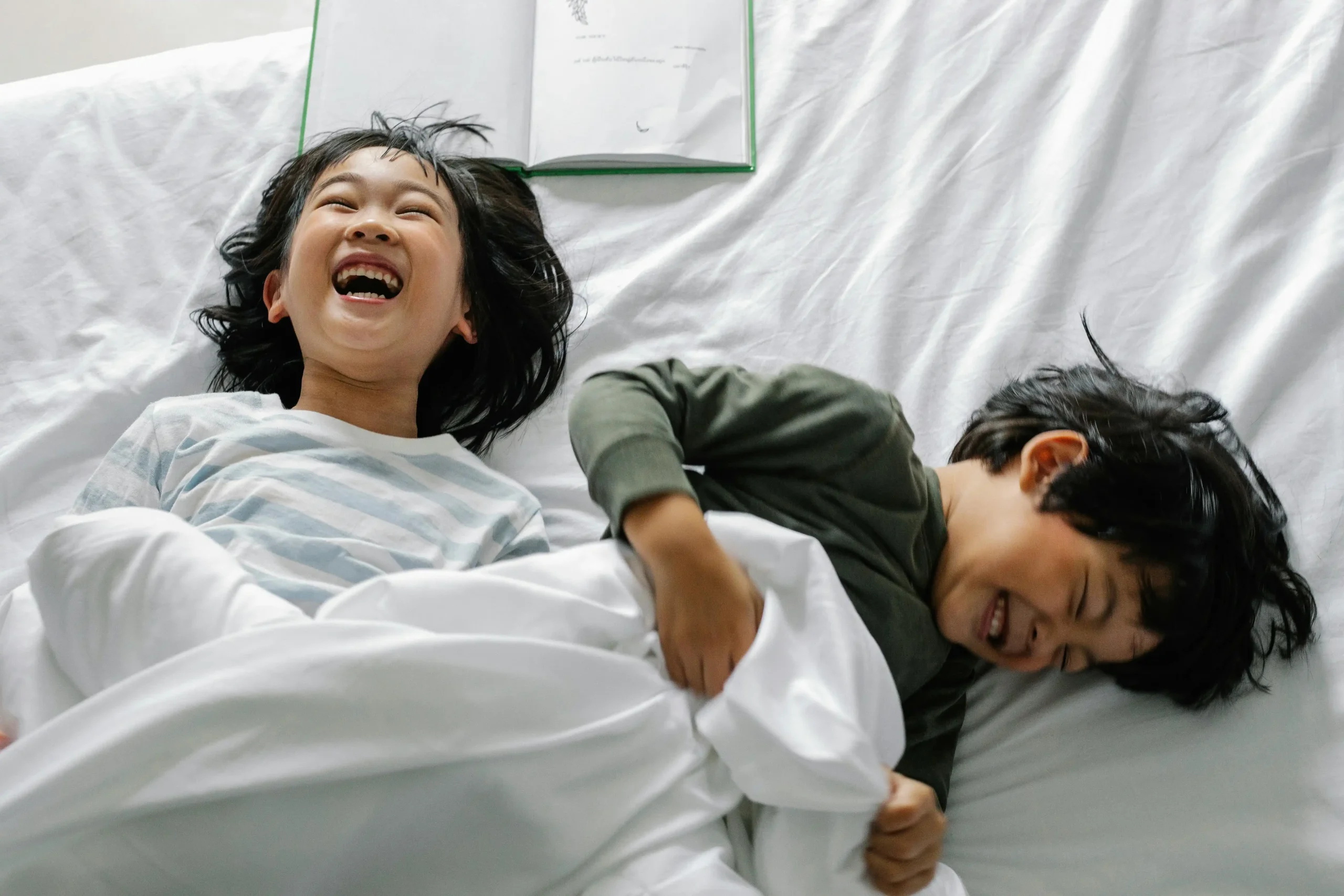 Two children laughing and playing under a blanket while lying on a bed