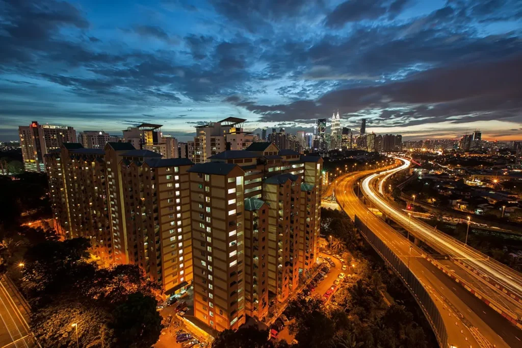 Long exposure photography of highway traffic light trails passing high-density apartment blocks in Kuala Lumpur at night.