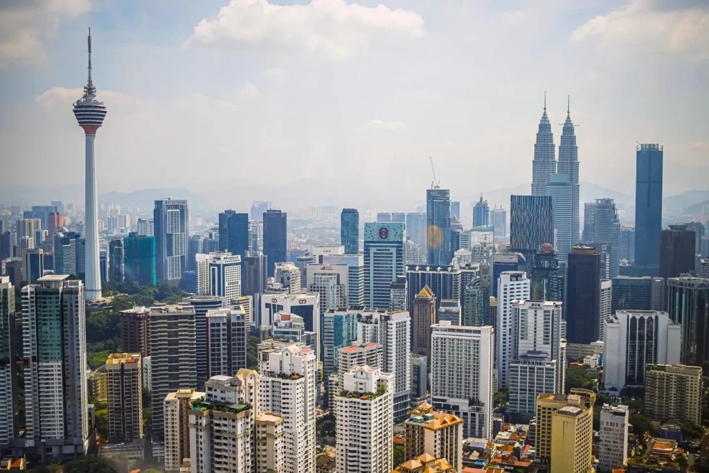 Panoramic daytime view of Kuala Lumpur city skyline featuring the KL Tower (Menara KL) and Petronas Twin Towers under a clear blue sky.
