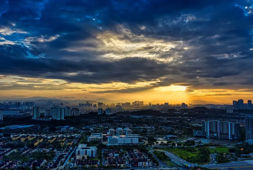 Dramatic sunset with sun rays breaking through dark clouds over a sprawling Malaysian residential township and city horizon.