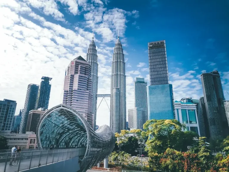 A low-angle view of the Petronas Twin Towers and modern glass skyscrapers in Kuala Lumpur against a blue sky.