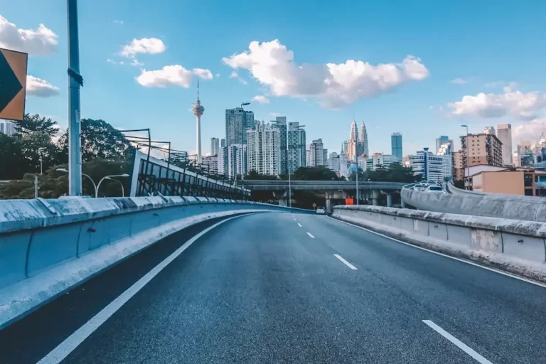 A view from a highway road looking towards the Kuala Lumpur skyline with KL Tower visible in the distance, under a blue sky.